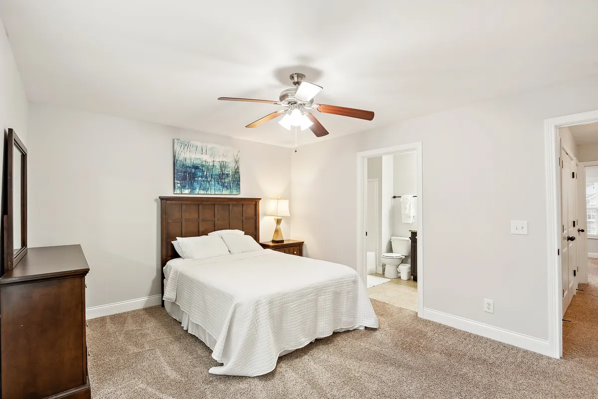 This image displays a serene bedroom featuring a comfortable bed, nightstand, tasteful artwork, dresser, and a clear view of the doorway leading to an ensuite bathroom. Soft morning light fills the room, creating a welcoming and restful atmosphere.