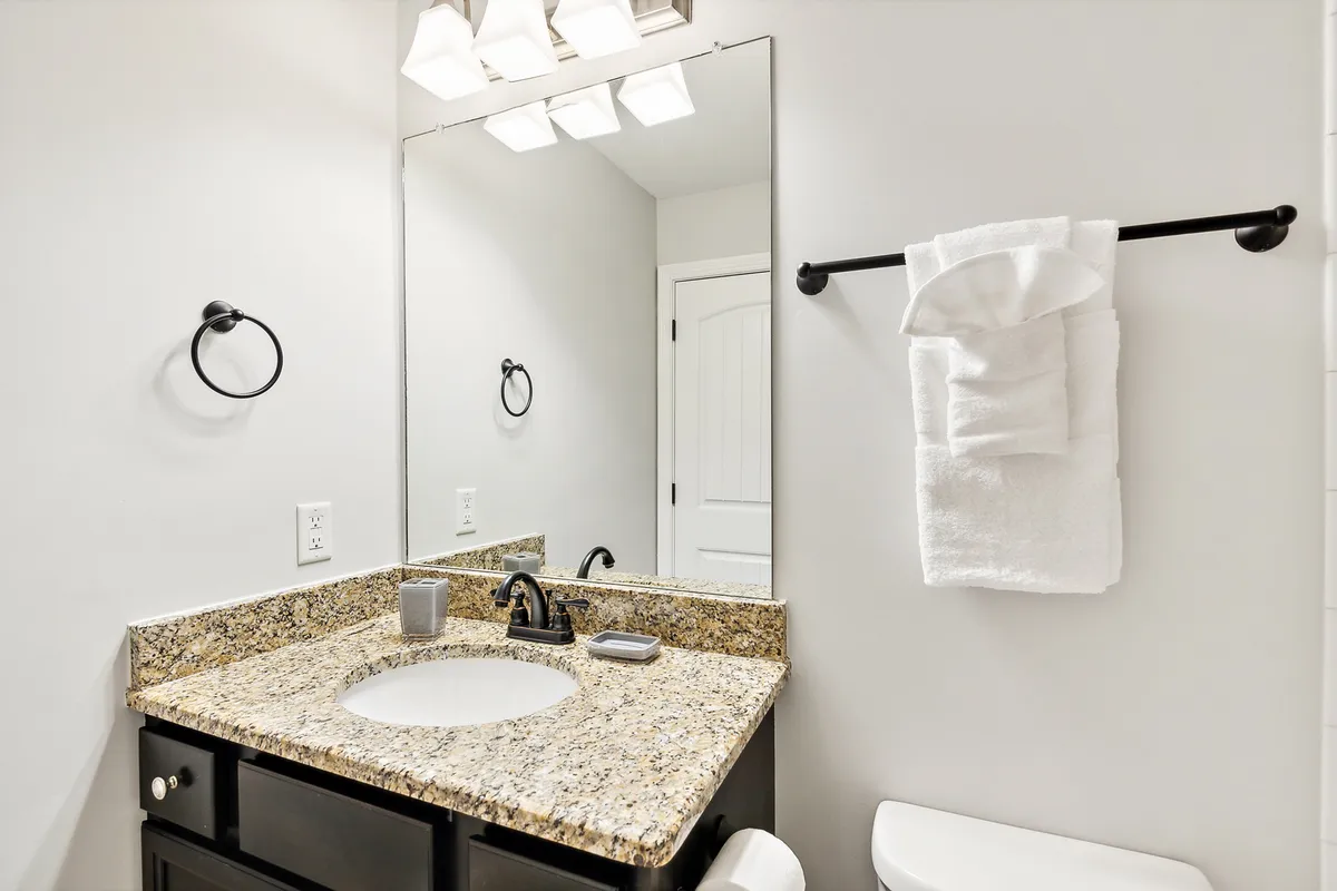 A fresh and inviting ensuite bathroom featuring a granite countertop, large mirror, sink, and elegant light fixtures. The space is designed for personal grooming and relaxation, with a clean, modern feel.
