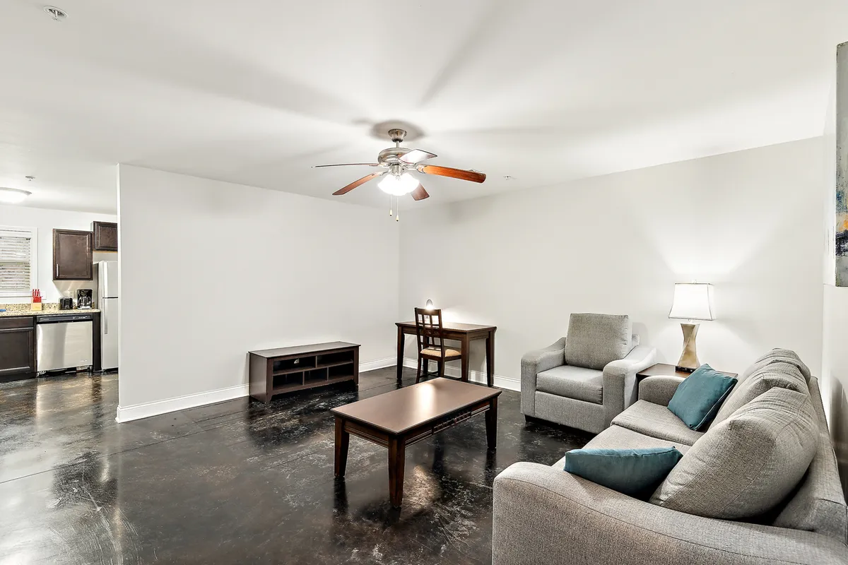 A bright, open living and dining area furnished with a comfortable sofa, a stylish coffee table, and a nearby dining table. Soft lighting from a contemporary lamp sets a welcoming tone, and the space is arranged to invite relaxation, conversation, or entertaining guests.